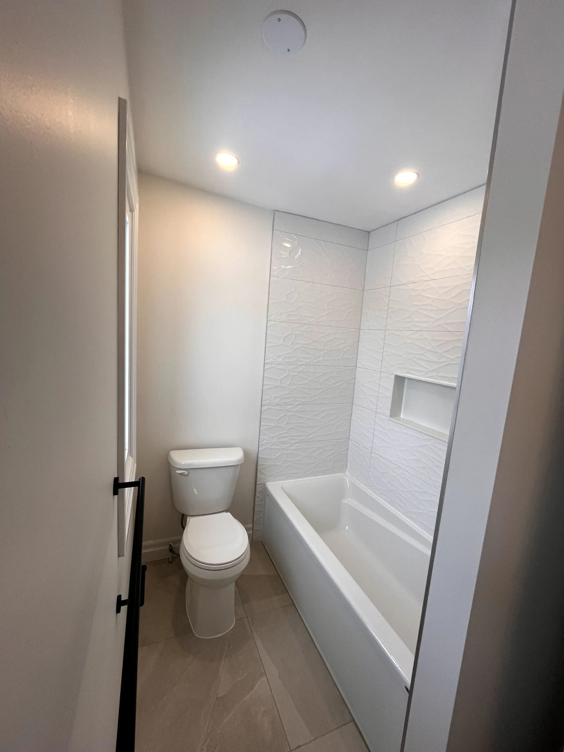 How to plan a bathroom renovation - Modern minimalist bathroom featuring a built-in bathtub, textured white tile shower wall with niche, recessed lighting, and large format floor tiles in a neutral tone