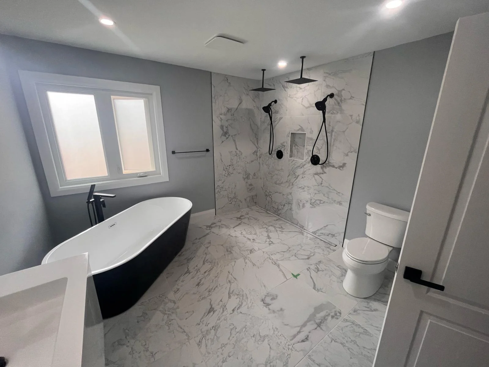 Modern bathroom renovation featuring a freestanding black and white bathtub, large walk-in curbless shower with dual rainfall showerheads, marble-look tiles, and a wall-mounted toilet—part of a complete 3-week remodel in the Greater Toronto Area.